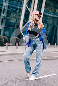 Young sexy blonde hipster woman posing on the street. Wearing blue stylish jacket, jeans and baseball hat and sunglasses