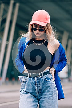 Young sexy blonde hipster woman posing on the street. Wearing blue stylish jacket, jeans and baseball hat and sunglasses