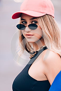 Young sexy blonde hipster woman posing on the street. Wearing blue stylish jacket, jeans and baseball hat and sunglasses