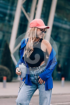 Young sexy blonde hipster woman posing on the street. Wearing blue stylish jacket, jeans and baseball hat and sunglasses