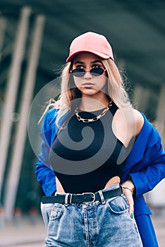 Young sexy blonde hipster woman posing on the street. Wearing blue stylish jacket, jeans and baseball hat and sunglasses