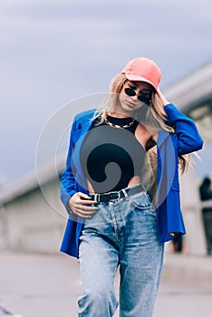 Young sexy blonde hipster woman posing on the street. Wearing blue stylish jacket, jeans and baseball hat and sunglasses