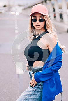 Young sexy blonde hipster woman posing on the street. Wearing blue stylish jacket, jeans and baseball hat and sunglasses