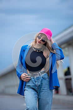 Young sexy blonde hipster woman posing on the street. Wearing blue stylish jacket, jeans and baseball hat and sunglasses