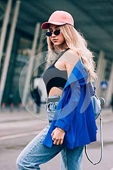 Young sexy blonde hipster woman posing on the street. Wearing blue stylish jacket, jeans and baseball hat and sunglasses