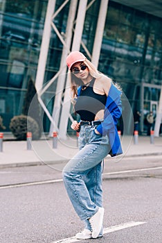 Young sexy blonde hipster woman posing on the street. Wearing blue stylish jacket, jeans and baseball hat and sunglasses