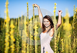 Young sexy brunette woman in white dress standing with eyes closed and enjoying sunshine in high grass on summer day