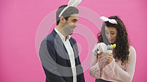 Young sexy couple on pink background. With hackneyed ears on the head. During this man gives a soft toy hare and colored