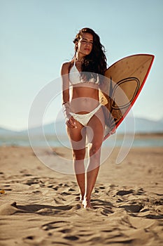 Young sexy female surfer is enjoying the sun while holding a surfboard and walking the beach at sea. Summer, vacation, sea