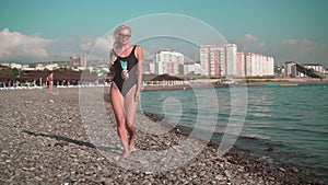 Young sexy girl in a bathing suit, walking the beach by the sea. Blonde in stylish sunglasses and a one-piece swimsuit.