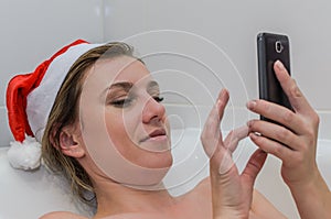 Young sexy girl in the bathroom in a New Year`s red Santa Claus hat communicates by video communication on a smartphone congratula