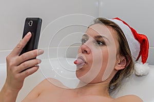 Young sexy girl in the bathroom in a New Year`s red Santa Claus hat communicates by video communication on a smartphone congratula