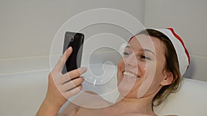 Young sexy girl in the bathroom in a New Year`s red Santa Claus hat communicates by video communication on a smartphone congratula