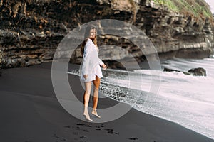 Young sexy girl in white shirt and swimsuit resting on black sand beach.