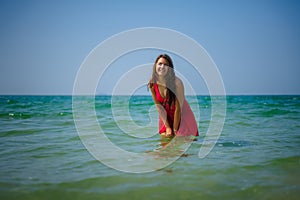 Young sexy long-haired brunette in red beach dress stands in the turquoise water of the ocean on a hot day. Beautiful girl