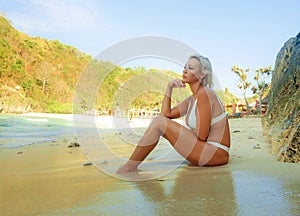 Young sexy and relaxed blond woman in bikini sitting on the sand looking at sea enjoying summer holidays at beautiful tropical