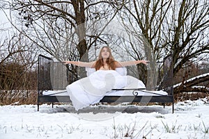 young, sexy, seductive, redhead woman sitting in the cold snowy winter nature in bed, arms on the iron bedstead