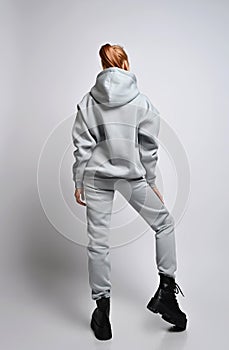 Young sexy slim red-haired woman stands back to camera demonstrating grey hoodie, pants and black massive shoes