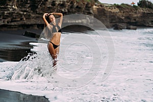 Young sexy sporty girl in black swimsuit walking on black sand beach.