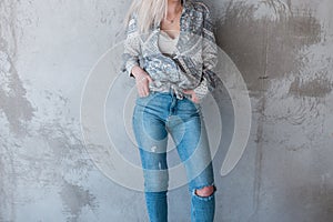 Young sexy stylish woman in a summer fashionable shirt in ripped stylish jeans is standing near a gray concrete wall.