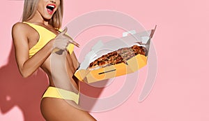 Young sexy tanned slim blond excited woman in sexy yellow beach bikini pointing at whole fresh pizza in box she holds