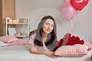 Young sexy woman on bed in pink pajamas with bouquet of red roses, room decorated with balloons. Postcard Happy Valentine`s Day,