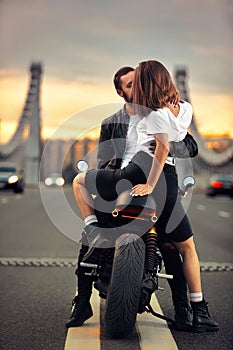 Young sexy woman hugging cute man in stylish black leather jacket, sitting on sports motorcycle on the bridge in the
