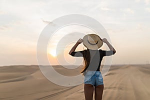 Young sexy woman with straw hat walking barefoot on desert dunes at sunset