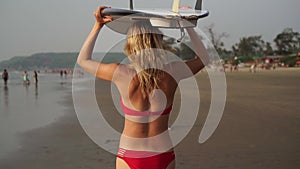 Young sexy woman surfer in a red swimsuit is walking along the beach with a surfboard