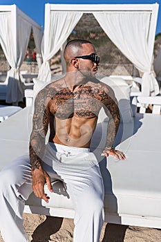 Young, shirtless man with tattoos sitting on a sunlounger posing for the camera
