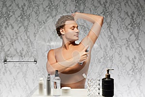 Young shirtless man use perspirant raising hand and applying it on underarm area against marbled wall in bathroom. photo