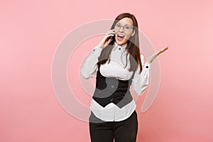 Young shocked amazed business woman in glasses holding talking on mobile phone spreading hands on pastel pink