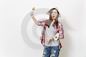 Young shocked beautiful woman in casual clothes and newspaper hat holding paint brush and wallpaper roll isolated on