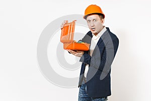Young shocked businessman in dark suit, protective hardhat holding opened case with instruments or toolbox isolated on