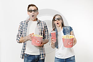 Young shocked couple, woman and man in 3d glasses watching movie film on date, holding bucket of popcorn, plastic cup of