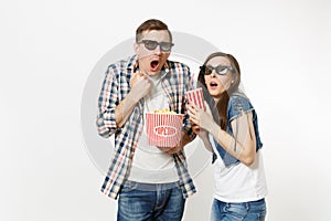 Young shocked couple, woman and man in 3d glasses watching movie film on date, holding bucket of popcorn and plastic cup