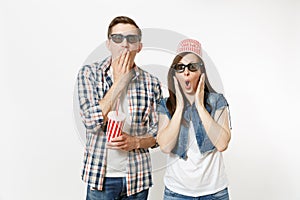 Young shocked couple, woman and man in 3d glasses with bucket for popcorn on head watching movie film on date, holding