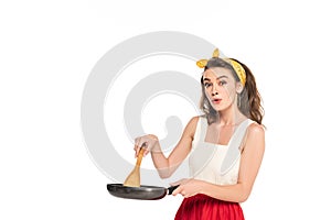 Shocked housewife in dress and apron holding frying pan and wooden spatula isolated on white