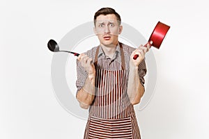 Young shocked man chef or waiter in striped brown apron, shirt holding red empty stewpan black ladle isolated on white