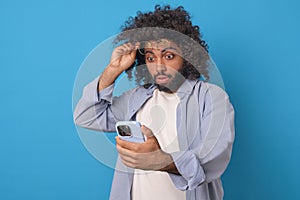 Young shocked optimistic Arabian man with phone lifts glasses from eyes