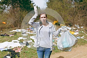 Young shocked woman in casual clothes, gloves cleaning holding trash bags and spreading hands in littered park. Problem