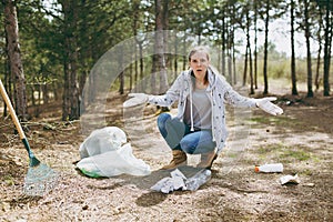 Young shocked woman in casual clothes and gloves cleaning rubbish and spreading hands near trash bags in park. Problem