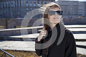 Young short haired girl in sunglasses and stylish black coat posing for portrait on grey urban city background