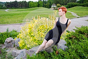 Young, short-haired, redhaired woman in a black dress sits on a stone in a park