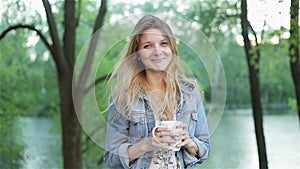 Young shy girl with long hair holds a cup of tea or coffee in hands against the blurred nature background. Pretty woman