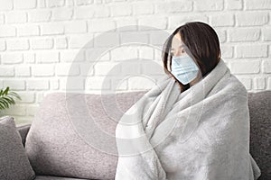 Young sick asian woman covered with blanket sits upset on sofa at home, wearing medical protective mask, looks away