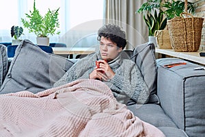Young sick male in sweater with mug of hot drink under plaid