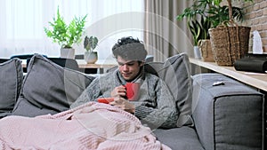 Young sick male in sweater with mug of hot drink under plaid relaxing using smartphone