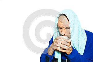 Young sick man have common cold flu and he is drinking a cup of hot tea while he is wearing towel on the head and thick winter clo