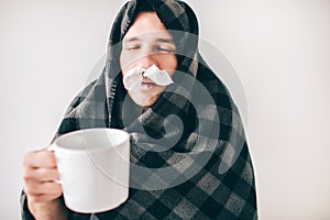 Young sick man isolated over white background. Guy covered with blanket. Holding cup of tea with closec nose tissues